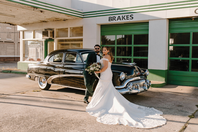 married couple emerald and gold standing in front of vintage car and garage