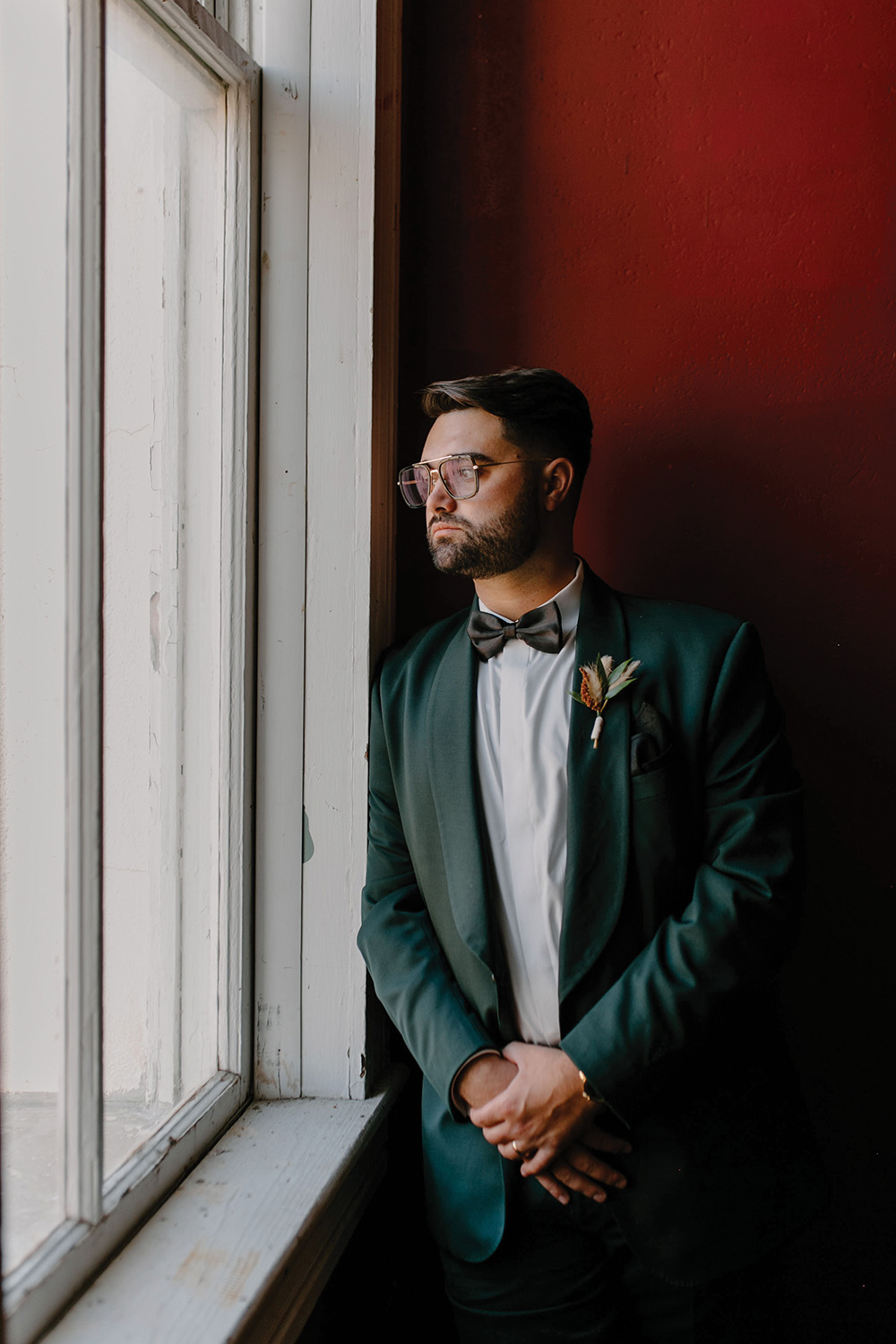 groom dark emerald green suit looking out a window in front of deep red wall
